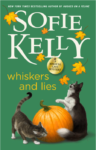 Whiskers and Lies by Sofie Kelly, A Magical Cats Mystery cover. Two cats playing around a pumpkin with leaves falling from above them over a green field.